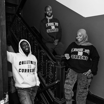 Three people on a brownstone staircase wearing Graetful Community Is Currency sweatshirts